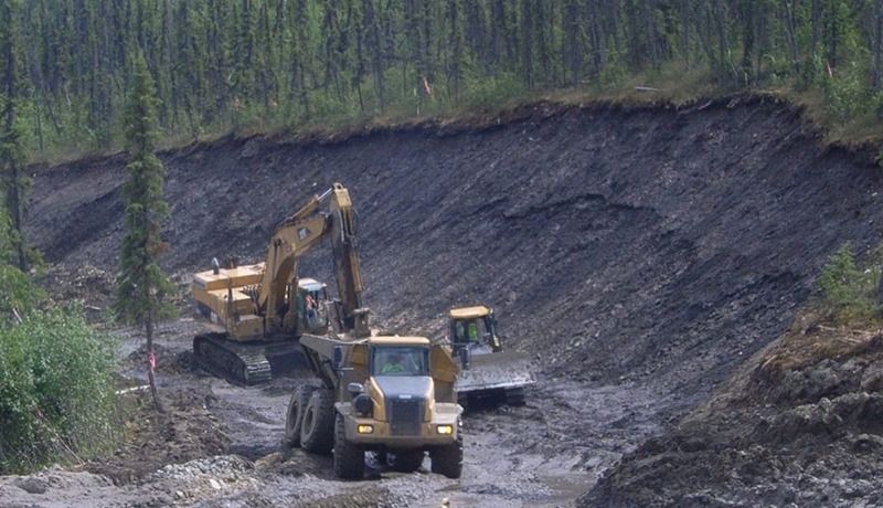 Construction vehicles, including an excavator, dump truck, and bulldozer, work on a muddy, partially forested site with a steep dirt embankment and trees in the background.