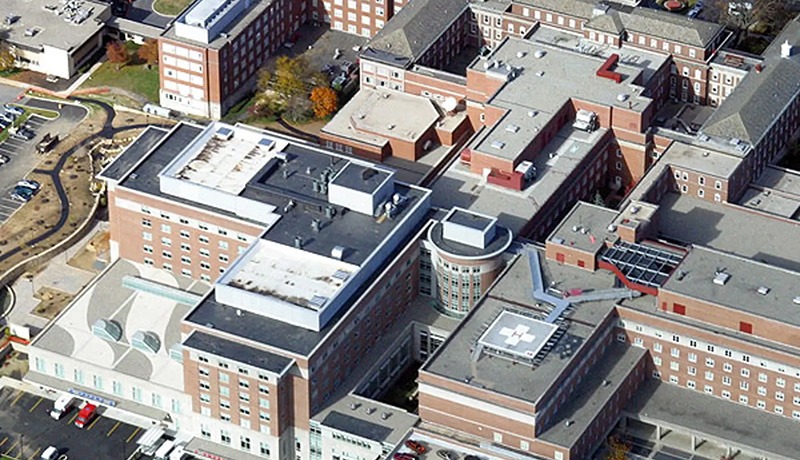 Aerial view of a large hospital complex with multiple interconnected brick buildings and flat roofs, surrounded by parking lots and roads. Some trees with autumn foliage are visible.