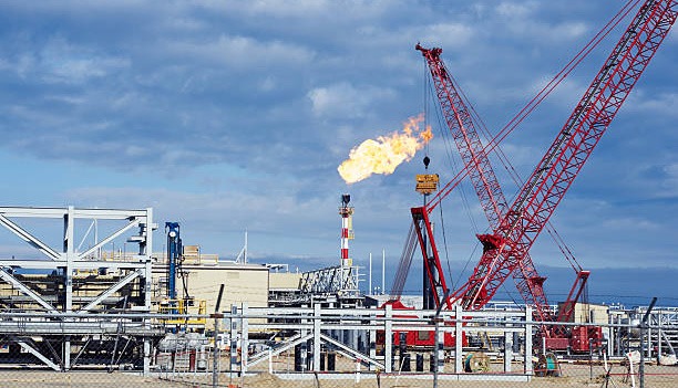 A large industrial site with a red crane, metal structures, and a tall smokestack emitting fire, under a partly cloudy sky. The scene is enclosed by a metal fence.