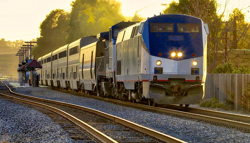 A passenger train with a blue and white engine travels along curved tracks at sunrise, passing through a small rural station surrounded by trees and fences.
