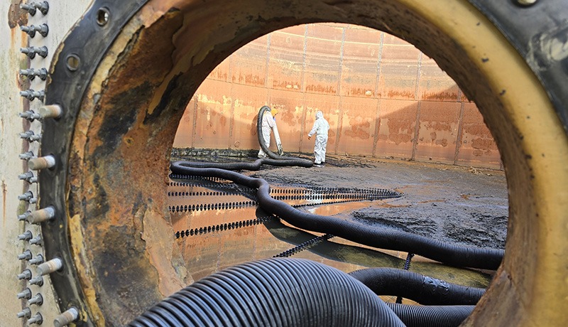 Two workers in protective suits are seen through a large circular opening, inside an industrial tank with large hoses and muddy residue on the floor. The tank walls appear rusty and stained.