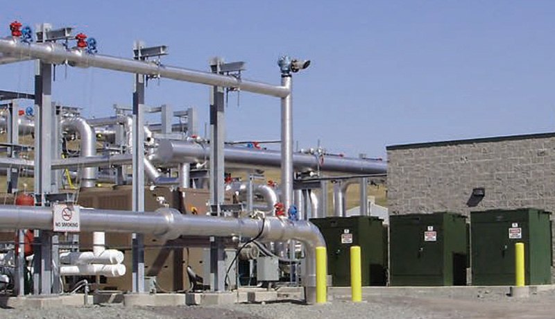 Industrial facility with large metal pipes, valves, and utility boxes outdoors on gravel ground next to a building with a cinder block wall; clear sky in the background.