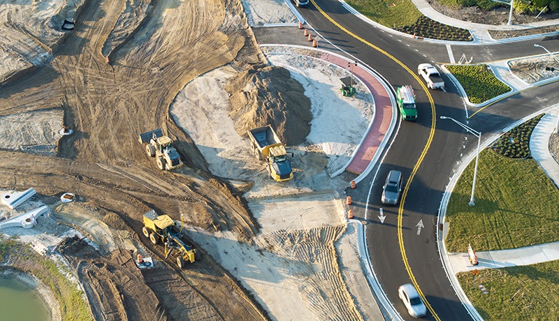 Aerial view of a construction site near a road with several vehicles and construction equipment moving dirt. Cars are driving on the newly paved curved roads beside the work area.