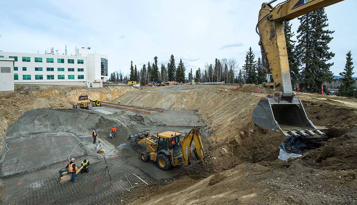 Construction site with heavy machinery and workers. An excavator is digging near the edge, with backhoes and a bulldozer at the base. Several workers wearing safety vests stand on the site, with a white building in the background.