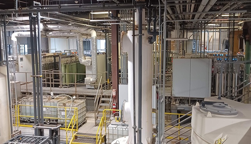 An industrial facility interior with numerous pipes, tanks, and metal walkways. Various control panels, storage containers, and insulated equipment are visible, suggesting a manufacturing or chemical processing environment.