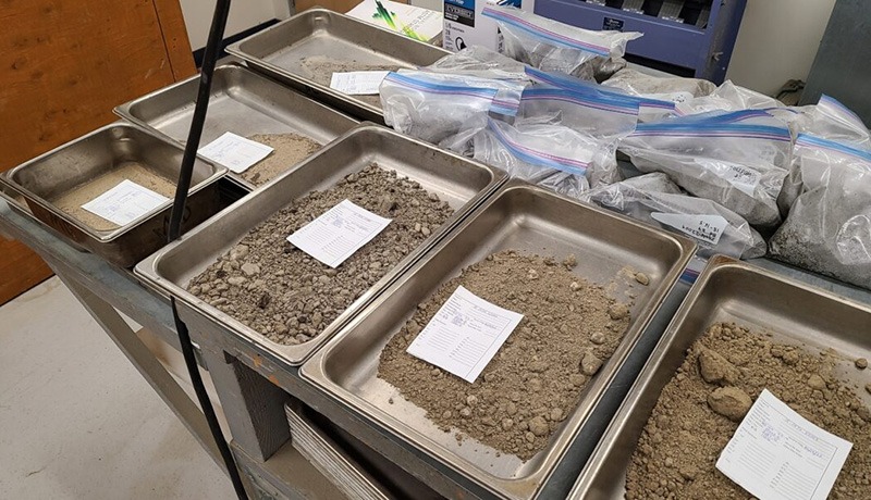 Several metal trays contain different soil samples with paperwork on top. In the background, plastic bags filled with more soil samples are visible on a laboratory cart.