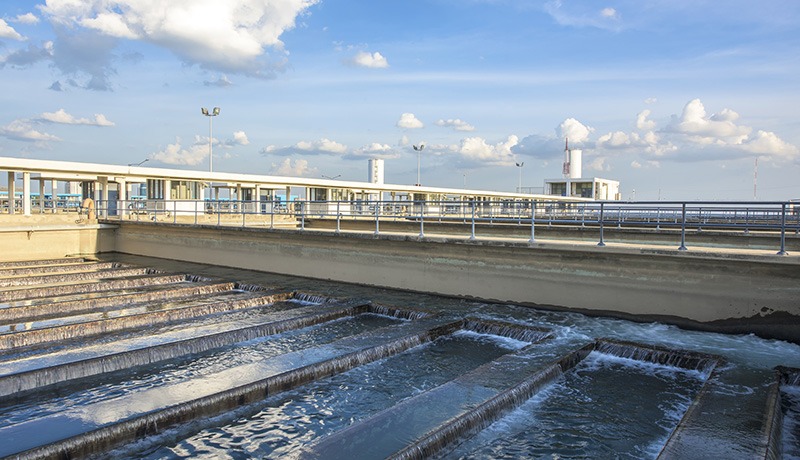 A modern water treatment facility with cascading water flowing through multiple rectangular channels, metal railings, and structures under a blue sky with scattered clouds.