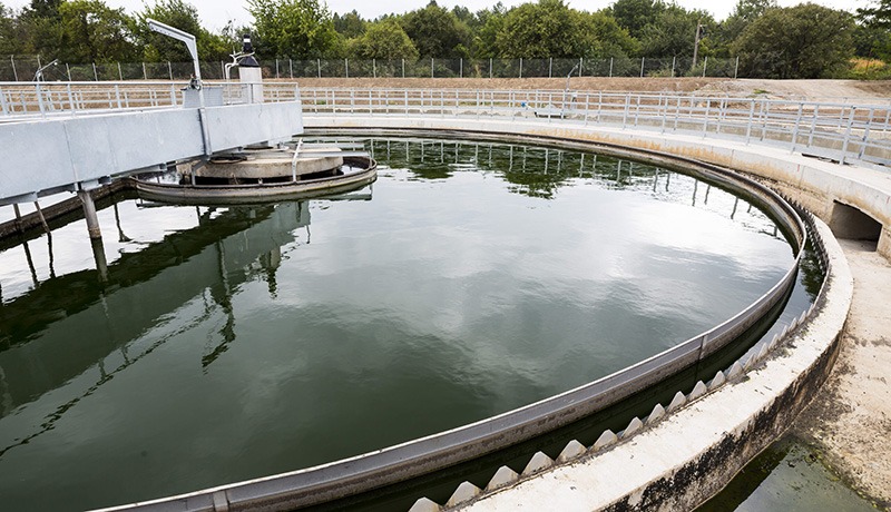 A large circular water treatment tank at a wastewater treatment plant, surrounded by metal railings and trees in the background. The water inside the tank appears still and reflective.