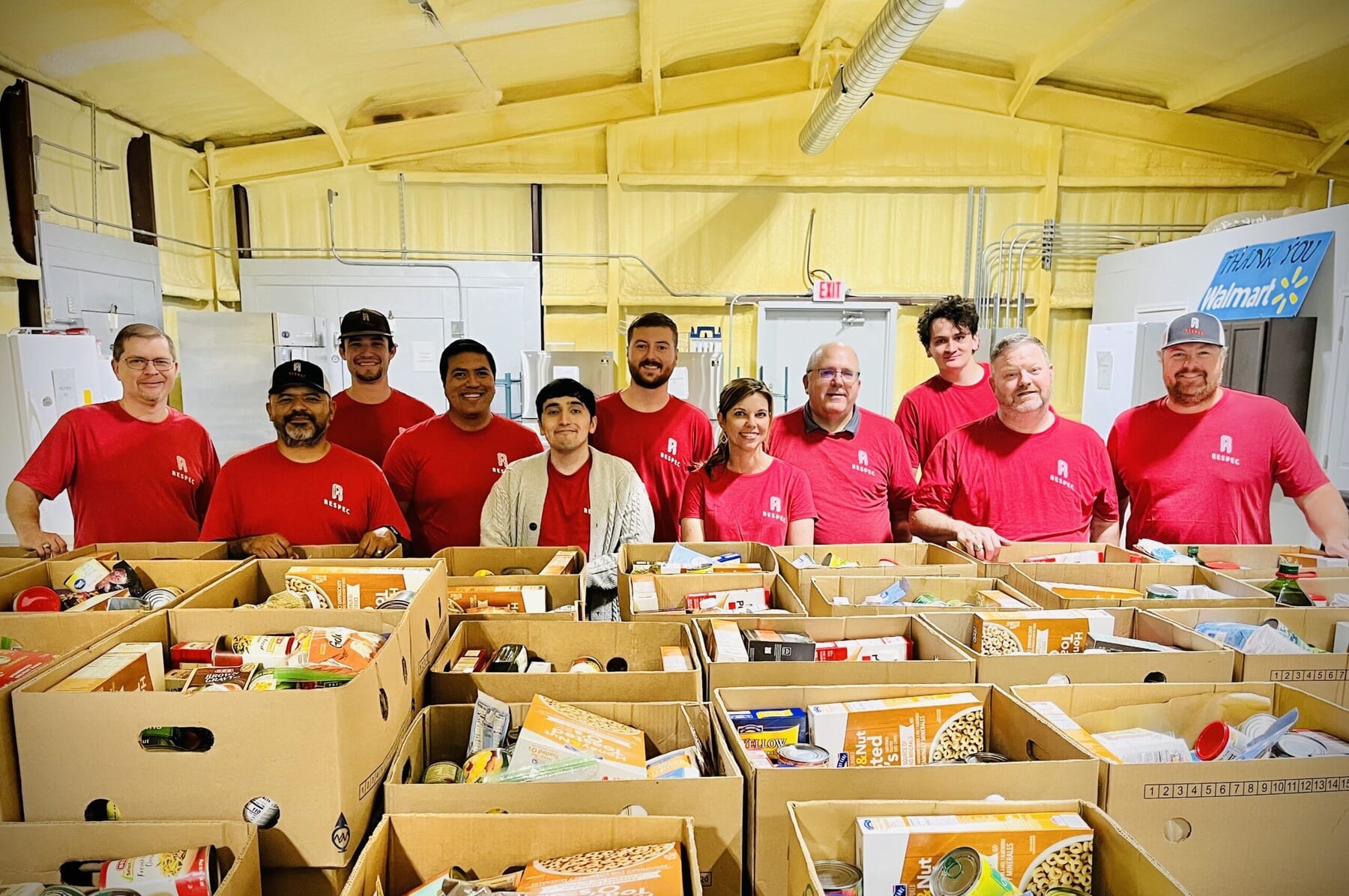 A group of volunteers wearing matching red shirts stands behind rows of boxes filled with assorted food items inside a food bank. They are smiling and posing for the photo.