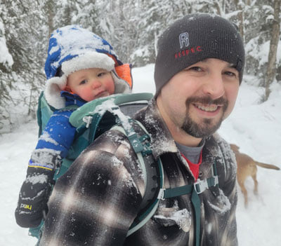 A man smiles while carrying a bundled-up baby in a backpack carrier on a snowy forest trail. Snow covers their clothing and trees, and a dog is visible in the background.
