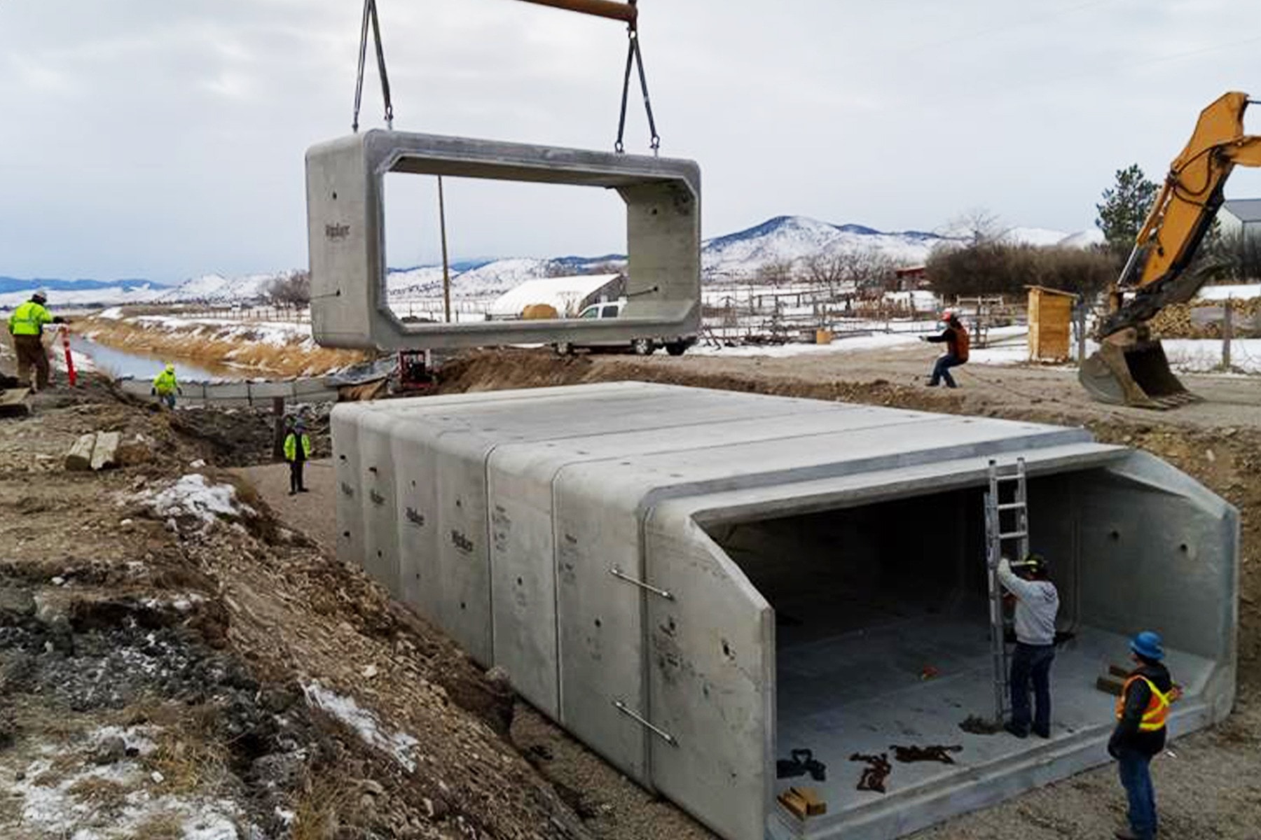 Construction workers guide a large concrete box culvert segment being lowered by a crane onto another segment in an open trench, with snowy mountains and construction equipment in the background.