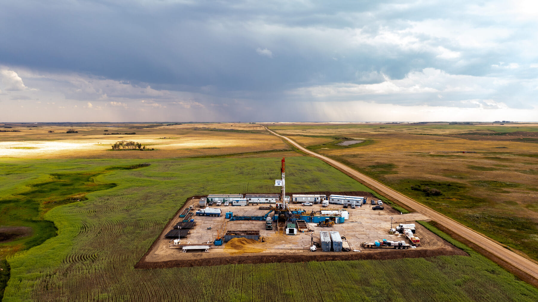 Aerial view of an oil drilling site surrounded by vast green fields under a partly cloudy sky, with a long dirt road stretching into the distance and rain visible on the horizon.