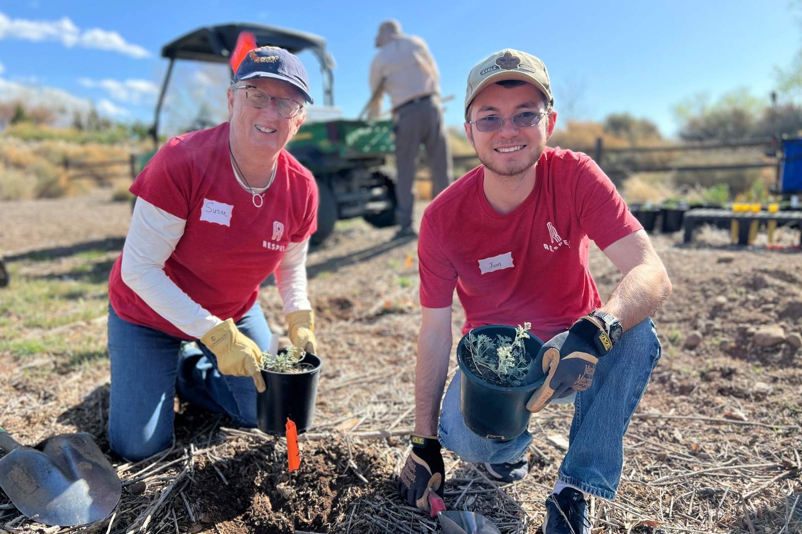 Two people wearing red shirts and gloves kneel outdoors, smiling while planting small plants in soil. They each hold a potted plant. A person and a green utility vehicle are visible in the background.