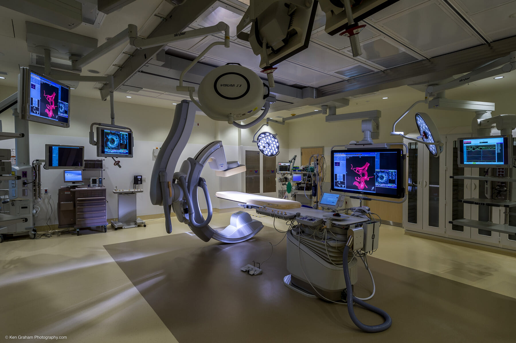 A modern hospital operating room features advanced medical equipment, monitors displaying medical images, overhead surgical lights, and a central operating table, all arranged in a clean, organized environment.