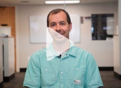 A man in a teal button-up shirt is smiling and sitting in an office with white walls and a whiteboard behind him. A transparent play button overlay appears on the image, suggesting it is a video thumbnail.