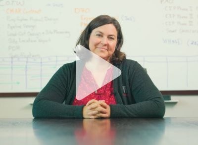 A woman with brown hair, wearing a red blouse and dark cardigan, sits at a table with her hands folded. A whiteboard with text and diagrams is in the background, and a video play icon is overlaid on the image.