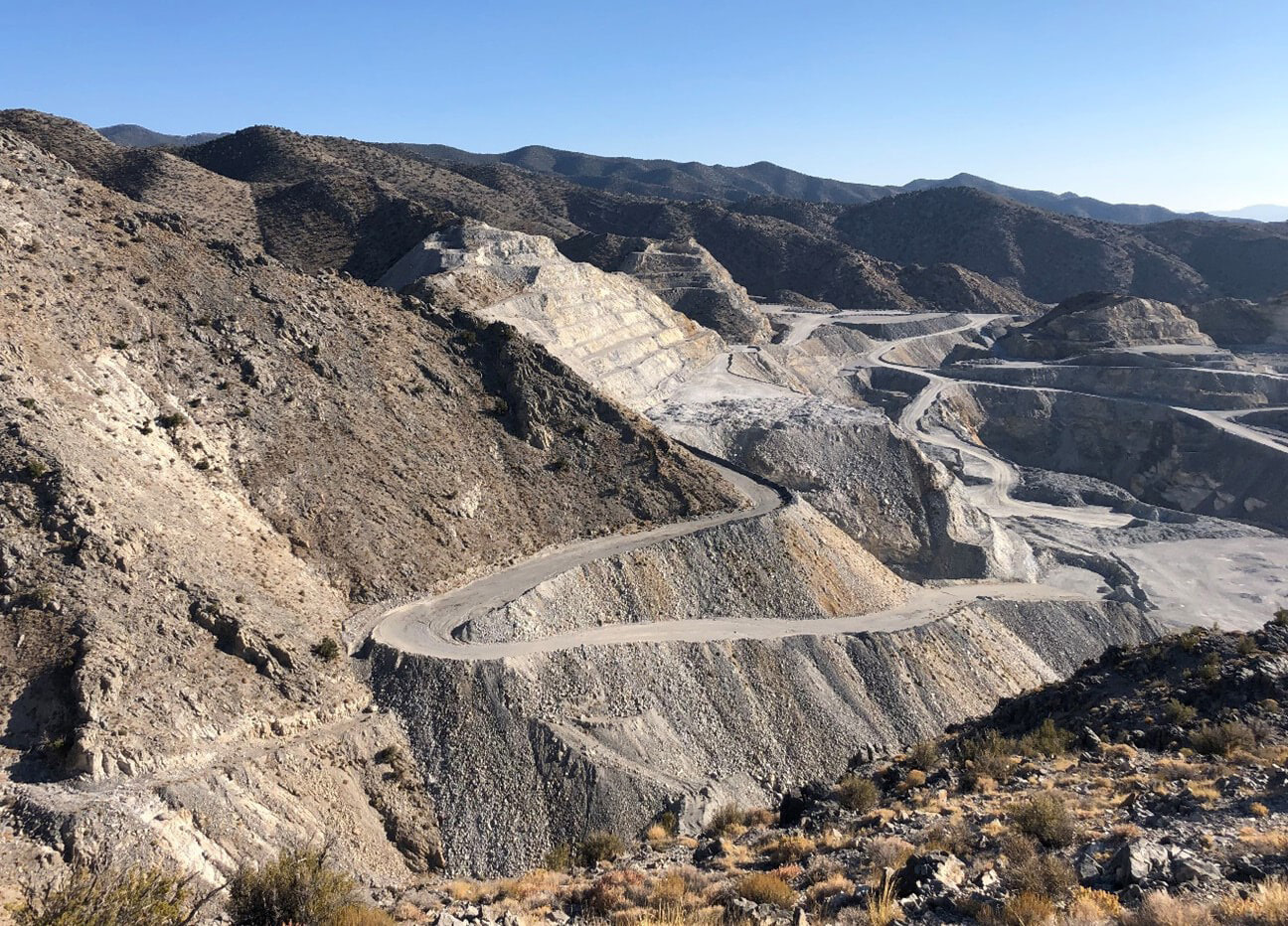 An open-pit mine with terraced slopes and winding roads is set in a dry, mountainous landscape under a clear blue sky. Sparse vegetation is visible in the foreground.