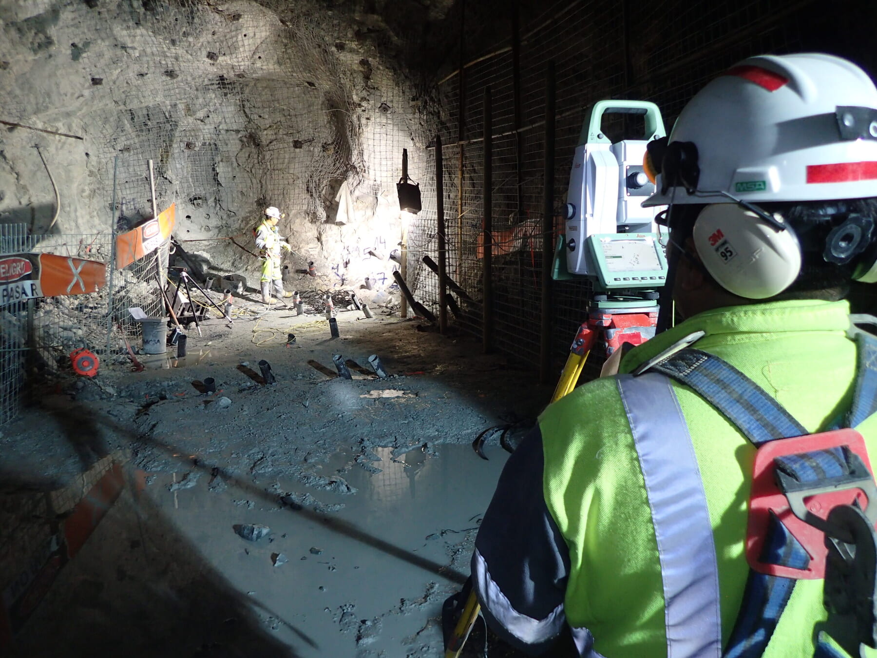 A worker in safety gear operates surveying equipment inside an underground tunnel, while another worker stands further ahead near construction materials and tools, illuminated by artificial light.