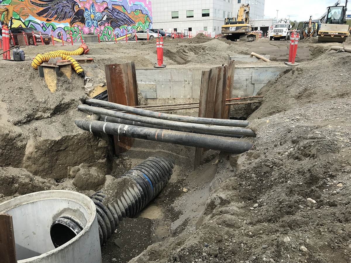 Excavated construction site with exposed pipes and large drainage hose, surrounded by dirt, metal supports, safety barriers, and machinery; a mural on a nearby building is visible in the background.