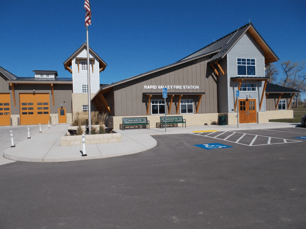The image shows the Rapid Valley Fire Station, a modern building with tan and gray siding, orange doors, a tall tower, flagpole, and accessible parking spaces in front under a clear blue sky.