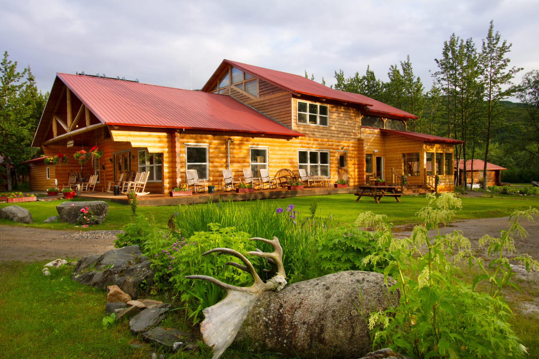 A large wooden lodge with a red roof sits surrounded by green grass, trees, and colorful flowers; antlers rest on a rock in the foreground, and hanging baskets decorate the porch under a partly cloudy sky.