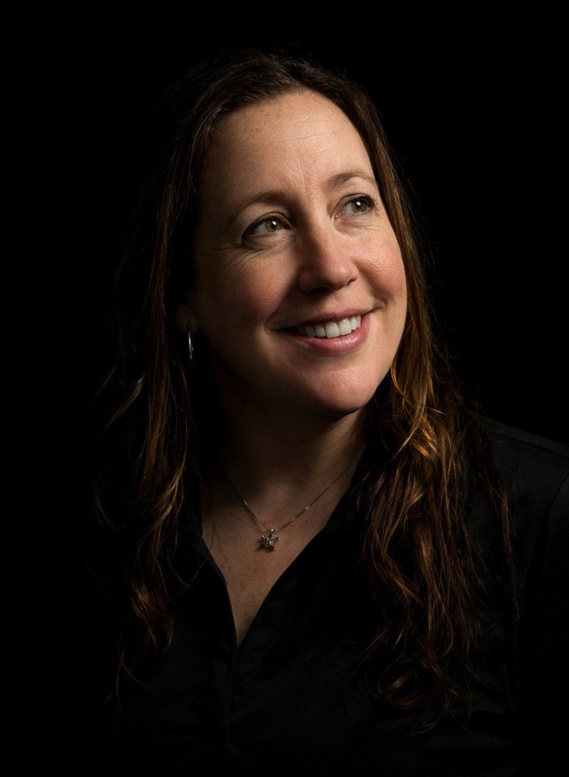 A woman with long brown hair smiles while looking up and to the side. She is wearing a black top and a necklace with a star-shaped pendant. The background is dark, and soft lighting highlights her face.