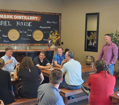 A group of people sit around wooden tables in a distillery tasting room, listening to a man standing and speaking. A chalkboard with drink options and barrels decorates the background.