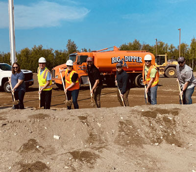 Seven people wearing construction gear and safety vests stand in a line holding shovels at a groundbreaking event, with a large orange Big Dipper construction truck and equipment in the background.