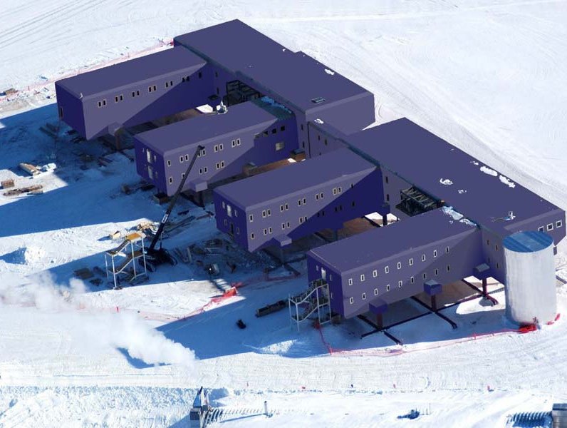 Aerial view of a modern, modular building with several connected purple units elevated above snowy ground in an Antarctic landscape, surrounded by snow, equipment, and structures.