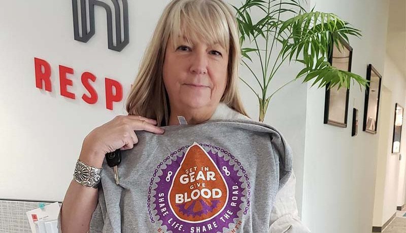 A woman with blonde hair holds up a T-shirt that says Get in Gear, Give Blood in an office with a plant and the word RESP on the wall behind her.