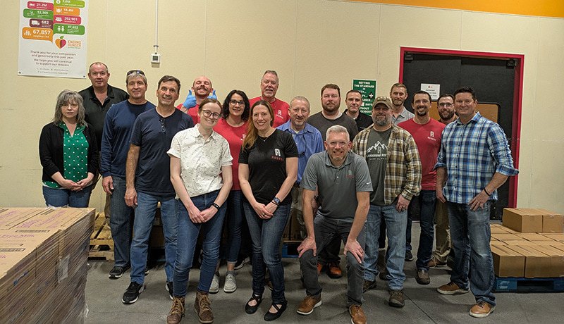 A group of 18 people poses for a photo inside a warehouse. They stand and kneel in front of stacks of boxes, smiling at the camera. The setting has industrial lighting, pallets, and colorful signage on the walls.