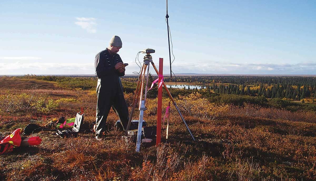 A person in outdoor gear stands beside scientific equipment on a tripod in a grassy, open landscape with trees and a lake in the background under a blue sky.