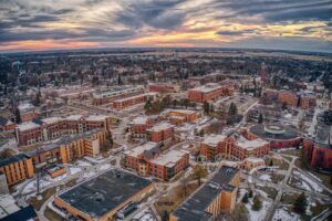 Aerial view of a university campus with multiple buildings, patches of snow on the ground, and a dramatic sunset sky in the background. The campus is surrounded by trees and a sprawling town.