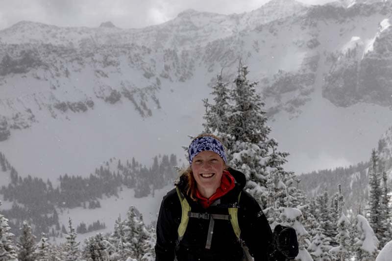 A person wearing outdoor gear and a patterned headband smiles while standing in a snowy mountain landscape with snow-covered trees and peaks in the background.