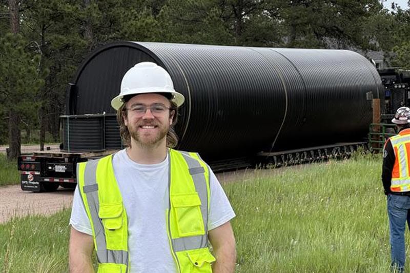 A man in a white hard hat and yellow safety vest smiles in front of a large cylindrical black structure being transported on a flatbed truck. Another worker is visible in the background. Trees line the area.