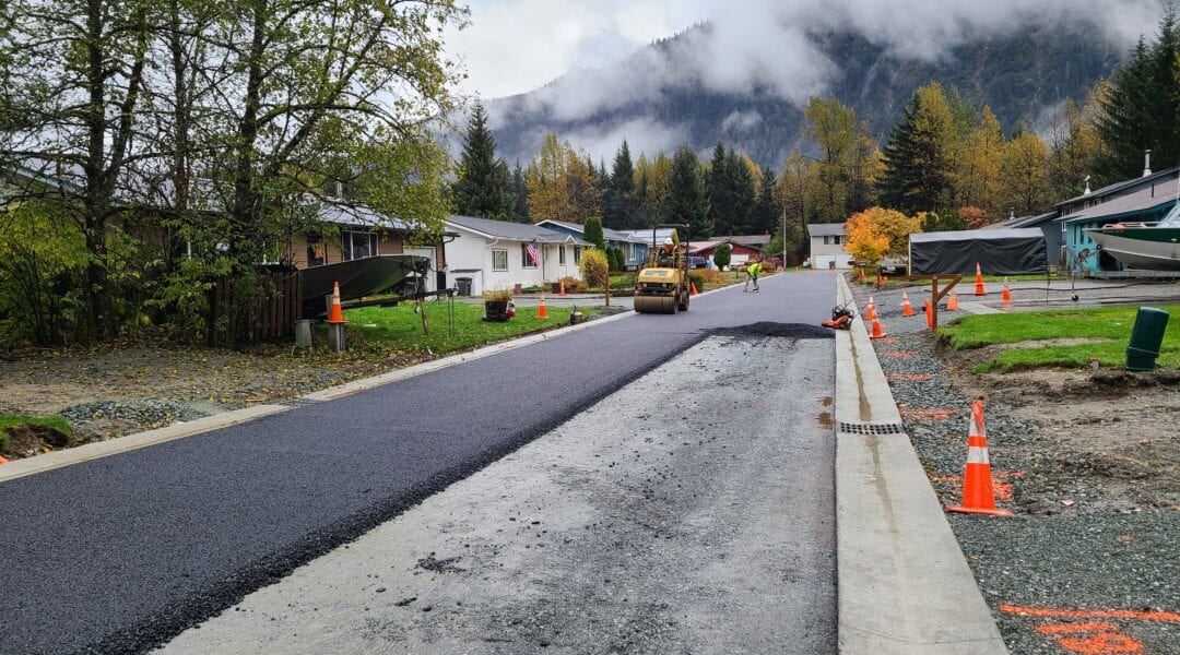 A freshly paved road in a residential neighborhood, with construction cones lining both sides. A steamroller is compacting asphalt; houses, trees, and misty mountains are visible in the background.