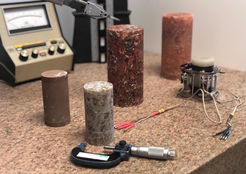 Several cylindrical rock samples sit on a countertop, surrounded by scientific instruments, including a caliper and an electronic device with wires and a gauge in the background.