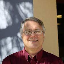 A middle-aged man with glasses and gray hair smiles, wearing a maroon collared shirt. He stands in front of a wall with sunlight and shadows creating patterns in the background.