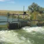 A small irrigation canal with a metal gate structure controls the water flow, surrounded by grassy fields and a solar panel on the right, under a clear blue sky.