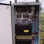 An open metal utility box contains batteries, circuit boards, fuses, and various colored wires. The box is mounted outdoors near a field, with numbered markings and a yellow label inside.