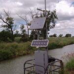 A weather monitoring station with two solar panels stands by a canal, surrounded by green grass and trees under a cloudy sky.