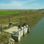 A small water control structure with metal gates and monitoring equipment sits by a canal, surrounded by grassy fields and farmland under a clear blue sky.