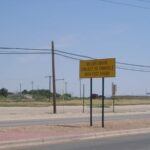 A yellow road sign reads, US 285 South Subject to Sinkhole 1000 Feet Ahead, standing beside a road with power lines and a flat, open landscape in the background.