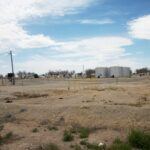 A barren, gravel-covered industrial site with scattered equipment, yellow railings, and large silver storage tanks in the background under a partly cloudy sky. Sparse vegetation grows in the foreground.