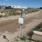 A solar-powered monitoring device is installed on a pole beside a mostly dry, muddy drainage canal, with industrial buildings and vehicles visible in the background under a blue sky.
