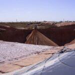 A large sinkhole with steep sides extends across a sandy, barren landscape. Construction workers and equipment are visible near the edge, highlighting the scale of the collapse. The sky is clear and blue.