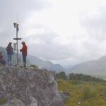Three people set up scientific equipment on a rocky outcrop overlooking a green valley with mountains in the background under a cloudy sky.