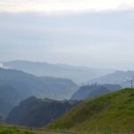 Rolling green hills in the foreground with layered, misty blue mountains fading into the distance under a cloudy sky. A small weather station stands on the grassy hill to the right.