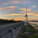 A rural road runs alongside a canal at sunset, with a metal monitoring station equipped with an antenna and solar panel standing near the water’s edge. Tall grass and clouds are visible under the colorful sky.
