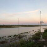 A canal runs alongside a grassy embankment at sunset. In the foreground, a metal weather or monitoring station stands near the water, with electrical wires and poles stretching into the distance.
